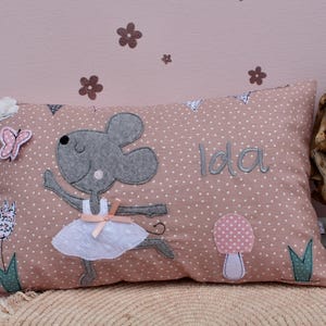 May include: Pink pillow with a gray mouse ballerina applique, a pink mushroom, and green leaves. The pillow has white polka dots and the name "Ida" embroidered on it.