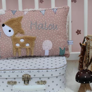 May include: A pink and brown pillow with a deer applique, white polka dots, and the name "Malou" embroidered on it. The pillow is sitting on top of a white suitcase with gray stars.