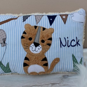 May include: A blue and white striped pillow with a brown and black tiger applique. The pillow has a white fluffy border and the name "Nick" embroidered on it.