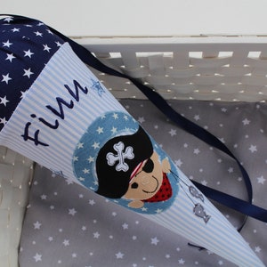 Könnte beinhalten: Eine blaue und weiße Schultüte mit Piratenmotiv. Die Tüte hat einen marineblauen oberen Teil mit weißen Sternen, hellblauen Streifen und dem gestickten Namen "Finn". Auf der Vorderseite befindet sich ein Pirat mit Augenklappe und Fischdesign.