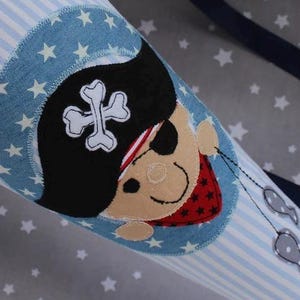 May include: A blue and white striped cone-shaped bag with a pirate design. The pirate has a black hat with a white skull and crossbones, an eye patch, and a red bandana. Two gray fish hang from the pirate's ear.
