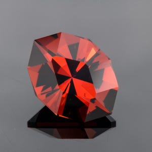 Op de afbeelding: Een gefacetteerde, dieprode edelsteen met een briljant slijpvorm, met scherpe geometrische hoeken en interne lichtreflecties. De steen heeft een levendige, verzadigde karmozijnrode tint met donkere, contrasterende schaduwen in de structuur.