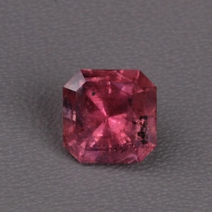 Könnte beinhalten: Ein facettierter, rechteckig geschliffener Edelstein in tiefem Rosa. Der Edelstein hat mehrere Facetten, die das Licht reflektieren und einen funkelnden Effekt erzeugen. Der Stein liegt vor einem grauen Hintergrund.