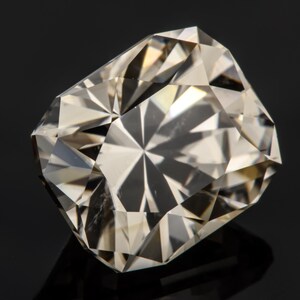 May include: A clear, faceted gemstone with a rectangular shape. The gemstone has a light brown hue and is set against a black background.