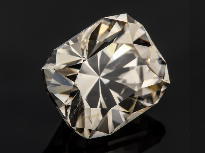 May include: A clear, faceted gemstone with a rectangular shape. The gemstone has a light brown hue and is set against a black background.