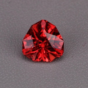 May include: A vibrant red gemstone cut into a triangular shape with multiple facets. The stone displays a brilliant, reflective surface with sharp edges and a deep crimson colour, showcasing a complex geometric pattern created by the precision cutting.