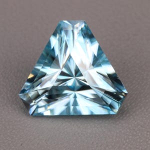 May include: A triangular-cut gemstone with a light blue color and a faceted surface. The stone features a complex geometric pattern of light and dark reflections across its top surface, creating a star-like effect radiating from the center.
