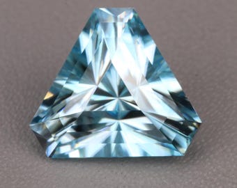 Può includere: Una pietra preziosa dal taglio triangolare di colore azzurro con una superficie sfaccettata. La pietra presenta un complesso motivo geometrico di riflessi chiari e scuri sulla superficie superiore, creando un effetto a stella radiante.