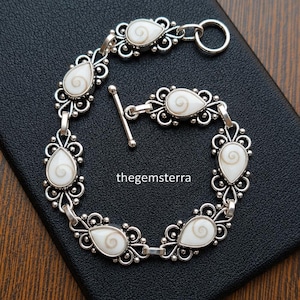 Pode incluir: Uma pulseira de prata com seis incrustações de concha olho de Shiva em forma de gota, inseridas em armações de metal com filigrana. A pulseira inclui um fecho de barra e o texto thegemsterra é visível no centro da disposição circular.