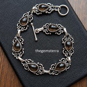 May include: A silver bracelet featuring six teardrop-shaped smoky quartz gemstones set in ornate metal filigree links. The bracelet includes a toggle clasp closure and displays the text thegemsterra in the center of the circular arrangement.