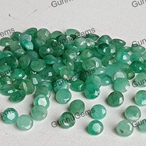 Op de afbeelding: Een verzameling ronde, gefacetteerde, groene smaragd edelstenen. De edelstenen liggen los op een wit oppervlak.