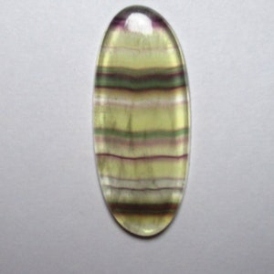 Puede incluir: Una piedra preciosa ovalada en cabuj&oacute;n con rayas verdes, moradas y blancas. La piedra preciosa est&aacute; tallada y pulida para la fabricaci&oacute;n de joyas.