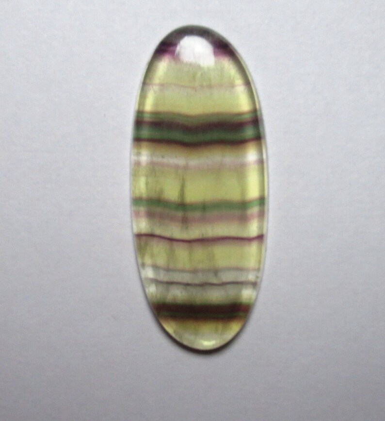 Puede incluir: Una piedra preciosa ovalada en cabuj&oacute;n con rayas verdes, moradas y blancas. La piedra preciosa est&aacute; tallada y pulida para la fabricaci&oacute;n de joyas.