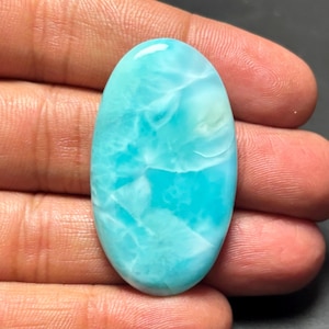 May include: Oval cabochon of blue and white larimar gemstone. The stone has a swirling pattern of blue and white.