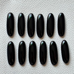 May include: Twelve black oval-shaped gemstones, likely onyx, arranged in two rows of six on a white surface.
