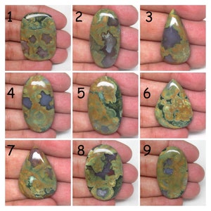 May include: Nine different green, brown, and purple color variations of a polished, oval-shaped, and teardrop-shaped gemstone. The gemstones are displayed on a hand.