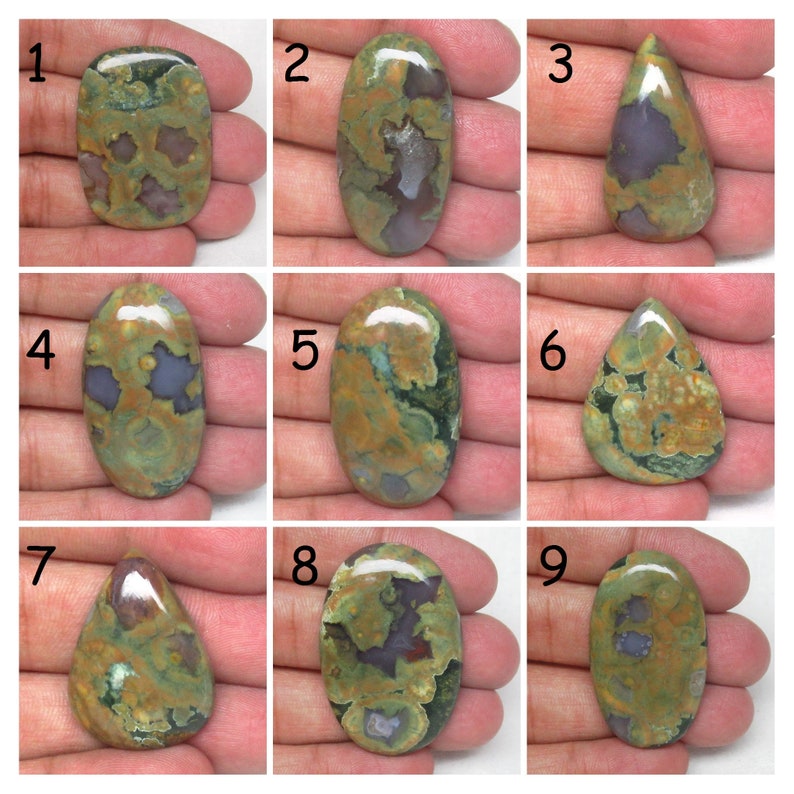 May include: Nine different green, brown, and purple color variations of a polished, oval-shaped, and teardrop-shaped gemstone. The gemstones are displayed on a hand.