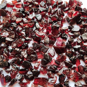 Op de afbeelding: Een verzameling ruwe, ruw geslepen granaat edelstenen. De stenen hebben een diepe, rijke rode kleur, met verschillende vormen en maten. De edelstenen zijn verspreid over het oppervlak.