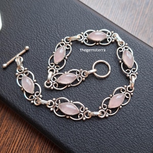 May include: A silver bracelet featuring six marquise-cut pale pink rose quartz gemstones set within intricate filigree metalwork. The bracelet includes a toggle clasp closure and the text thegemsterra is visible near the center of the piece.