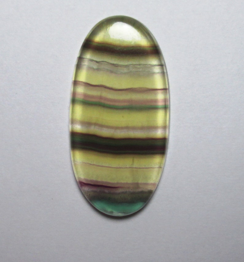 Puede incluir: Una piedra preciosa ovalada en cabuj&oacute;n con bandas verdes, amarillas y moradas. La piedra est&aacute; pulida y tiene un acabado liso y brillante.