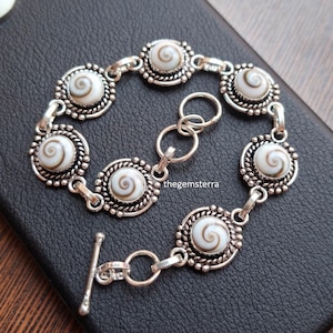 Peut inclure: Un bracelet argenté composé de six cabochons ronds en coquillage œil de Shiva, sertis dans une bordure en métal torsadé et perlé. Le bracelet est doté d'un fermoir à barre et porte l'inscription thegemsterra sur l'un de ses éléments.