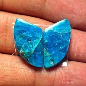 Op de afbeelding: Twee bijpassende, gepolijste blauwe edelstenen met een kristallijne structuur. De stenen zijn een levendig, doorschijnend blauw met gebieden van lichter turquoise en donkerdere insluitsels. Ze zijn bevestigd op een transparante lijmrug.