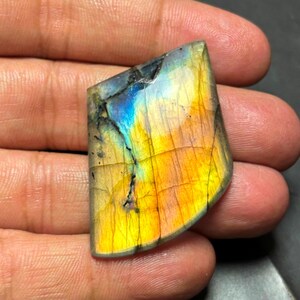 May include: A large, iridescent labradorite gemstone with a mix of yellow, orange, blue, and green colors. The stone is cut into a cabochon shape and has a smooth, polished surface.