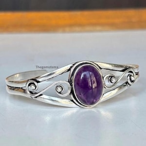 Peut inclure: Un bracelet manchette en argent orné d'une pierre précieuse améthyste violette de forme ovale au centre. La bande métallique présente des motifs de volutes complexes de chaque côté. Le texte Thegemsterra est visible sur le métal.