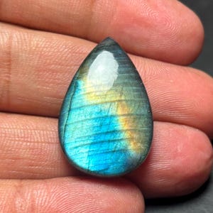 Puede incluir: Un cabujón de labradorita en forma de lágrima con un brillo iridiscente azul, verde y dorado.