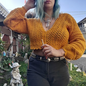 May include: Mustard yellow crochet top with a V-neck and long, flared sleeves. The cropped sweater has a scalloped hemline. The image shows a close-up of the top, highlighting the textured stitch pattern and the overall design.
