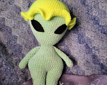 May include: Green crocheted alien plush toy with yellow crocheted hair. The toy is standing on a purple fabric with white drawings.