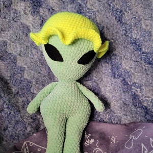 May include: Green crocheted alien plush toy with yellow crocheted hair. The toy is standing on a purple fabric with white drawings.
