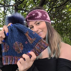 Puede incluir: Un gorro azul de crochet con un pompón y detalles coloridos. El gorro está siendo sostenido por una persona que lleva una diadema de crochet rosa.