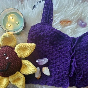 May include: Purple crochet top with a tie front and a delicate lace trim. The top is laid out on a white fluffy surface with a crocheted sunflower and crystals.