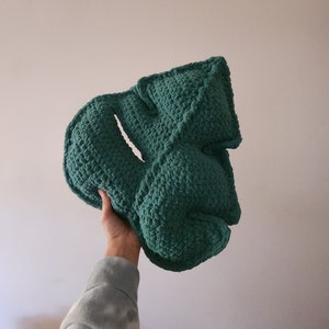 Puede incluir: Una almohada de forma de hoja hecha a crochet de color verde. La almohada está hecha de un hilo suave y texturizado y tiene una forma tridimensional.