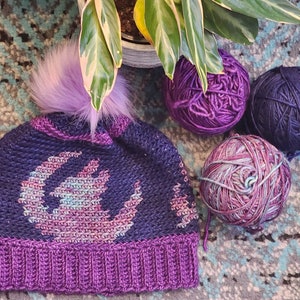 Puede incluir: Un gorro de punto hecho a mano, azul oscuro, con un borde morado y un gran pompón morado esponjoso. El gorro presenta un diseño en morado claro y rosa. Varias madejas de hilo en tonos morados y azules están junto al gorro.
