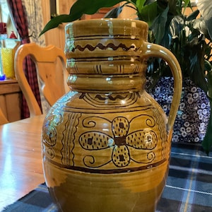 Puede incluir: Una jarra de cerámica marrón con una boca ancha y un asa. La jarra tiene un diseño decorativo de espirales y flores marrones y negras.