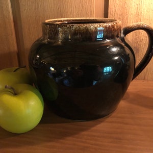 May include: A black ceramic pitcher with a brown glaze and a handle. The pitcher is sitting on a wooden surface with two green apples in front of it.