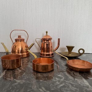 May include: A collection of antique copper kitchenware, including a kettle, a coffee pot, and several small pans. The copper items have brass handles and a warm, reddish-brown color. A small brass candlestick holder is also present.
