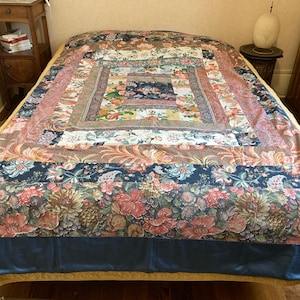 Peut inclure: Une courtepointe patchwork avec un motif floral dans des tons de rose, bleu et marron. La courtepointe a une bordure bleu foncé et une bordure dorée.