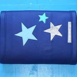 Könnte beinhalten: Eine dunkelblaue Lederclutch mit drei sternförmigen Verzierungen. Die Sterne sind in Blautönen und Silber gehalten. Die Clutch hat ein kleines Etikett mit der Aufschrift "Leatherware".