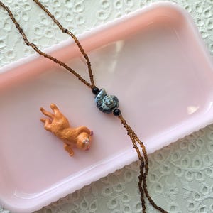 May include: A necklace featuring a long strand of small, translucent brown seed beads, a central blue and white swirled ammonite-shaped ceramic bead, and two small black spherical beads, accompanied by a small, orange, reclining cat figurine.