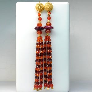 Könnte beinhalten: Ein Paar lange, baumelnde Ohrringe mit goldfarbenen Kugelsteckern. Die Ohrringe weisen eine Kombination aus roten, orangen und burgunderfarbenen Perlen auf, darunter runde und quadratische Formen. Das Design umfasst eine zentrale scheibenförmige Perle und eine quastenartige Anordnung von Perlen.