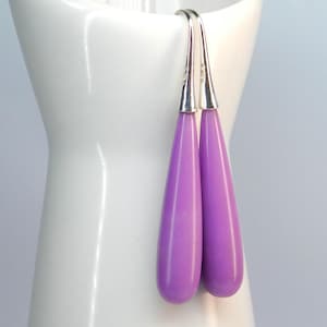 Op de afbeelding: Een paar lavendelkleurige druppelvormige oorbellen met zilverkleurige haken. De oorbellen hebben een gladde, glanzende afwerking en een eenvoudig, elegant ontwerp. De oorbellen worden gepresenteerd tegen een witte achtergrond.
