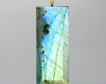 May include: A rectangular labradorite pendant with a gold-colored bail. The gemstone displays a mix of blue, green, and gray hues with visible internal lines. The pendant is set against a neutral background, highlighting its natural beauty.