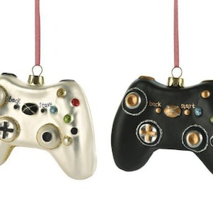 Op de afbeelding: Twee hangende ornamenten in de vorm van videogamecontrollers, één in glanzend wit en één in matzwart. Beide zijn voorzien van goudkleurige knoppen, een centraal rond embleem en de woorden "back" en "start" in kleine zwarte letters.