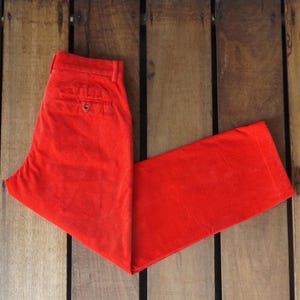 May include: Bright red corduroy pants, folded neatly on a wooden surface. The pants have a classic design with a button closure and pockets. The vibrant color and texture of the corduroy are clearly visible.