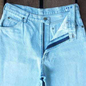May include: Light blue denim jeans with a partially unzipped fly. The jeans have classic five-pocket styling, a button closure, and a Wrangler label. The fabric appears faded, suggesting a vintage or worn look.