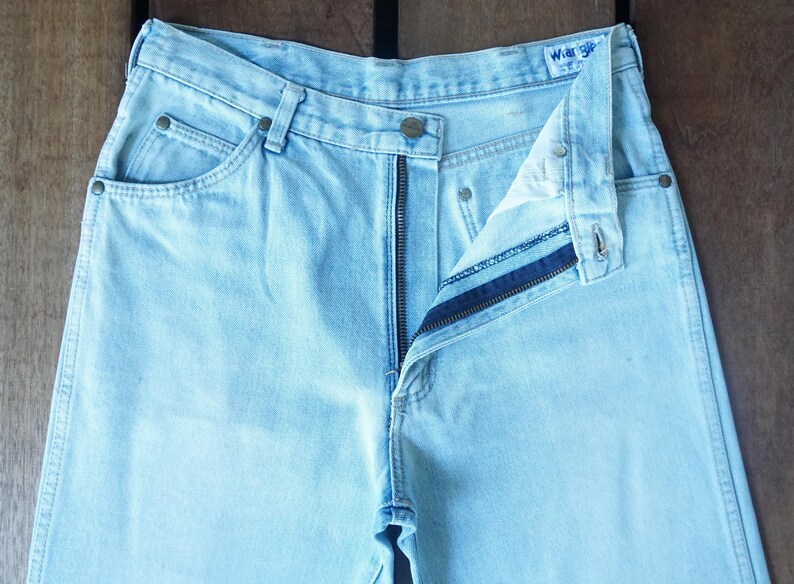 May include: Light blue denim jeans with a partially unzipped fly. The jeans have classic five-pocket styling, a button closure, and a Wrangler label. The fabric appears faded, suggesting a vintage or worn look.