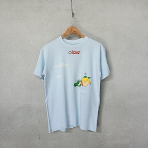 May include: Light blue t-shirt with a cartoon graphic of a blonde child wearing a green hat and a green vehicle. The text "The Jetsons" is printed in red on the top. The shirt is hanging on a wooden hanger.